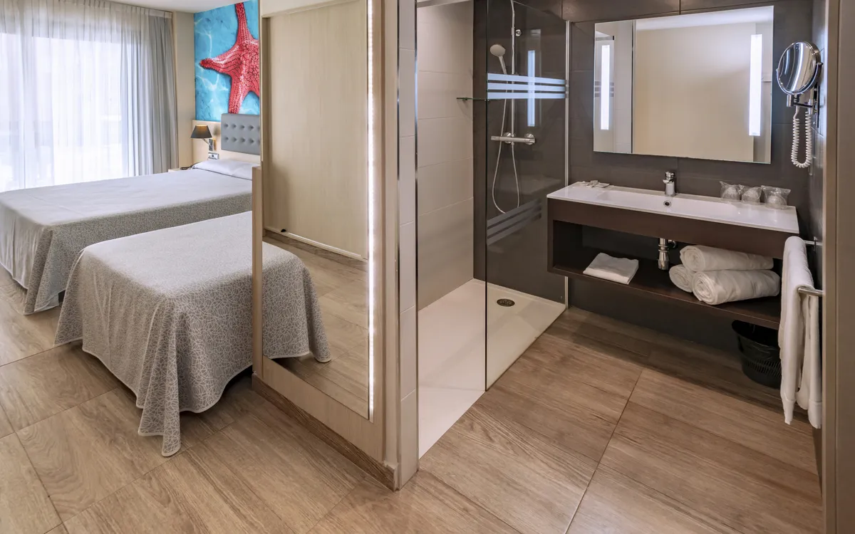 Man sieht im Hintergrund des modernen Doppelzimmers zwei Einzelbetten. Im Vordergrund istein sehr modernes und neues Duschbad zu sehen.